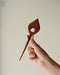 Elegant wooden hair clip shaped like a slender leaf, carved from durable teak wood with rich golden-brown grain, holding a loose chignon.