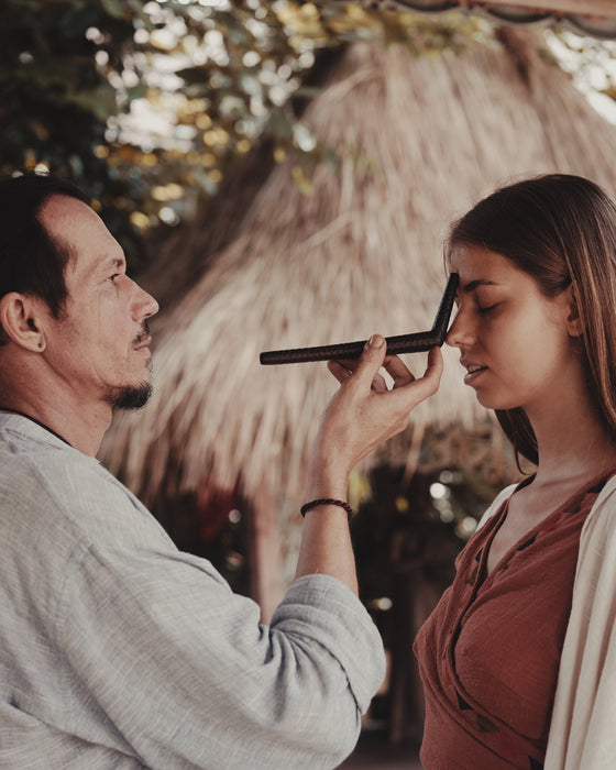 Person applying makeup to another person outdoors with a thatched roof in the background