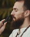 Man with a beard holding a black object close to his face against a blurred natural background