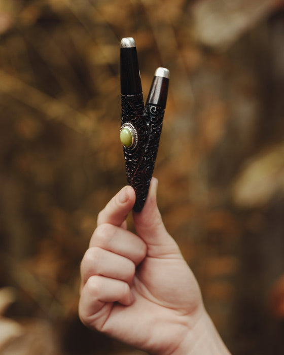 Hand holding a black and silver object with a green gemstone against a blurred natural background