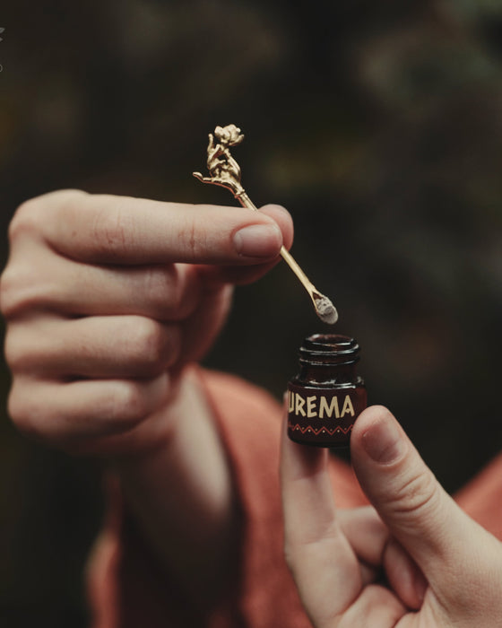 Person holding a small bottle labeled 'Burema' with a dropper and a dried flower.