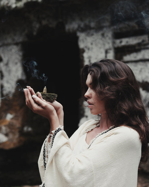 Woman holding a smoking incense bowl in front of a dark, textured wall.