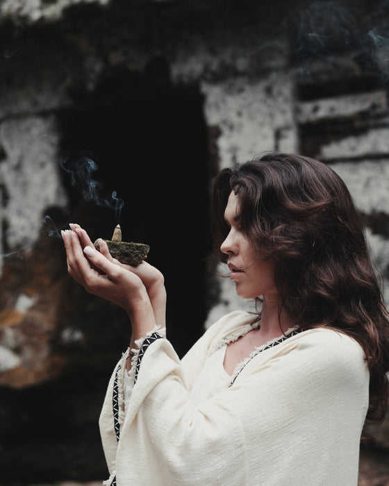 Woman holding a smoking incense bowl in front of a dark, textured wall.