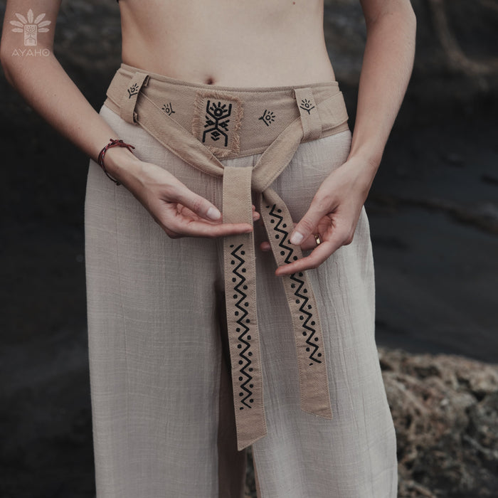 Person wearing a beige belt with black patterns, standing against a natural background.