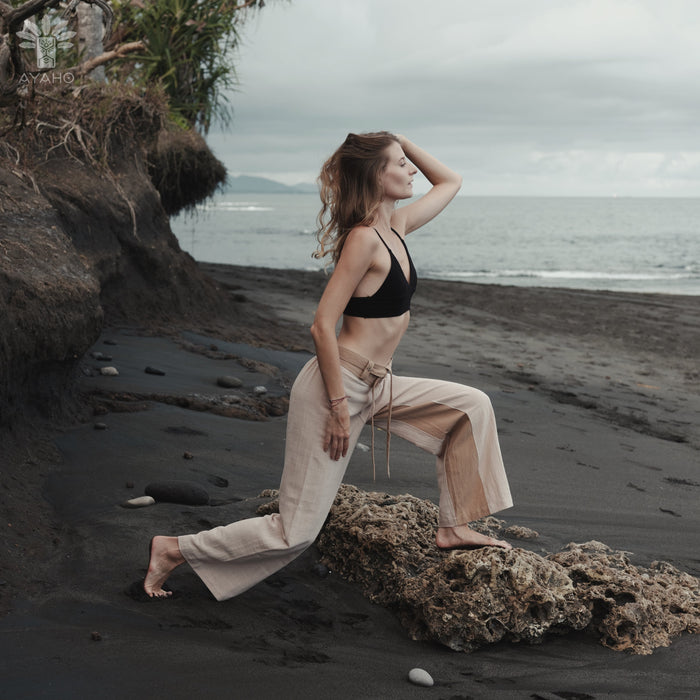 Woman in black bikini top and beige pants standing on a rocky beach with ocean and sky in the background