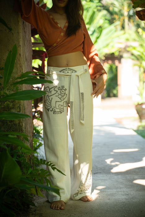 Person wearing a brown top and white pants with patterns, standing in a tropical setting.