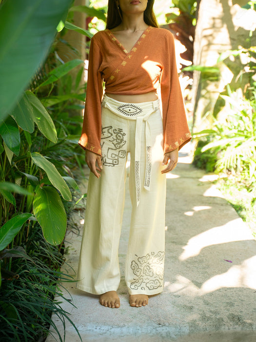 Person wearing a rust-colored top and white pants with patterns, standing in a garden.