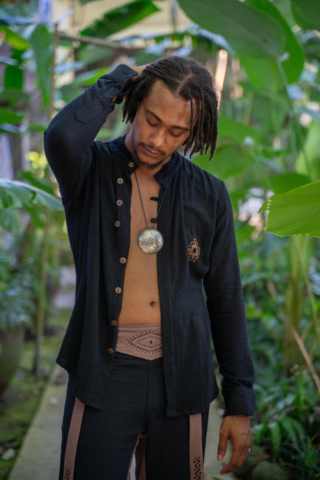 Man in a black jacket with buttons, beige shirt, and dark pants standing in a lush green outdoor setting.