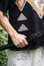 Person wearing a black garment with colorful geometric patterns, holding a black object against a blurred natural background.