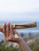 Hand holding a gold bracelet on a wooden stick with a blurred natural background