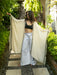 Woman holding a long, light-colored garment in an outdoor setting with greenery.