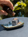 Hand pouring a liquid from a bottle onto a smoking cube on a small dish outdoors.