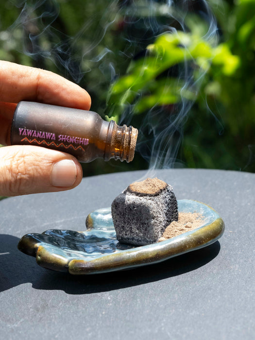 Hand holding a Yawanawa Shungite bottle over an incense burner with smoke rising.