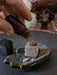 Hand pouring a liquid from a small bottle labeled 'Yawanawa Shungite' onto a stone in a decorative setting.