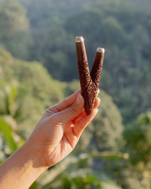 Hand holding two wooden slingshots with intricate designs against a blurred natural background