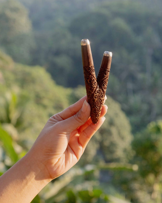Hand holding two wooden slingshots with intricate designs against a blurred natural background