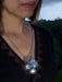 Close-up of a person wearing a necklace with a large, round, silver pendant against a blurred natural background.
