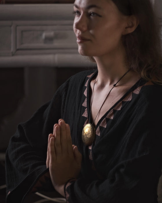Woman in a dark outfit with a gold necklace, hands clasped together in a dimly lit room.