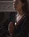 Woman in a dark outfit with a gold necklace, hands clasped together in a dimly lit room.
