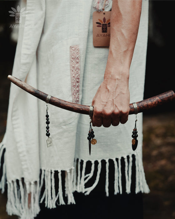 Person holding a curved wooden stick with decorative elements, wearing a white garment with fringe details.