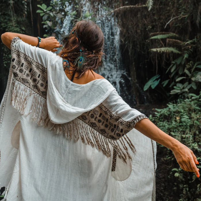 Woman in a white dress with a patterned shawl standing in front of a waterfall.