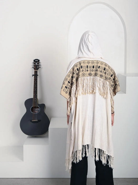 Person wearing a white shawl with intricate patterns and fringes, standing next to an acoustic guitar on a white surface.