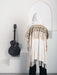 Person wearing a white shawl with intricate patterns and fringes, standing next to an acoustic guitar on a white surface.