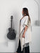 Woman wearing a white shawl with black patterns standing next to an acoustic guitar in a minimalistic setting.