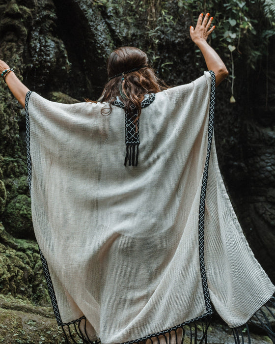 A person enveloped in a handmade Boho tribal cotton poncho, embracing the serenity of nature at a waterfall, reflecting a unisex shamanic clothing style