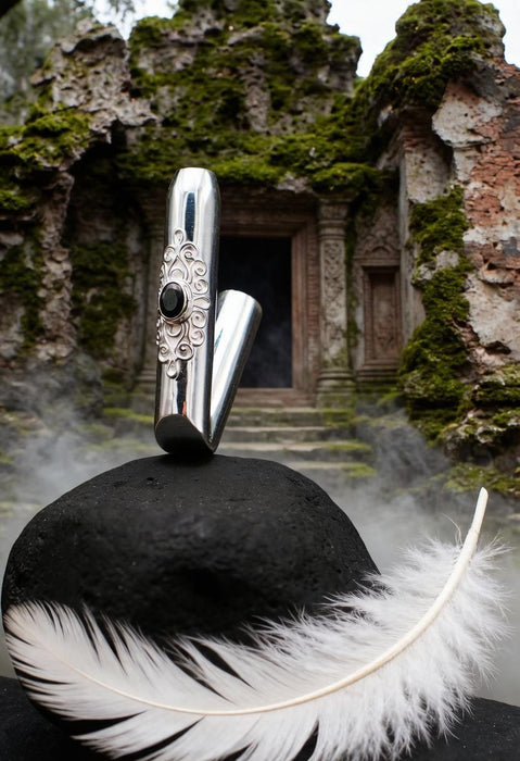 A polished silver metal object with ornate engraved detailing and a dark circular stone accent, balanced on a black surface with a white feather in the foreground and a mossy stone doorway behind it.